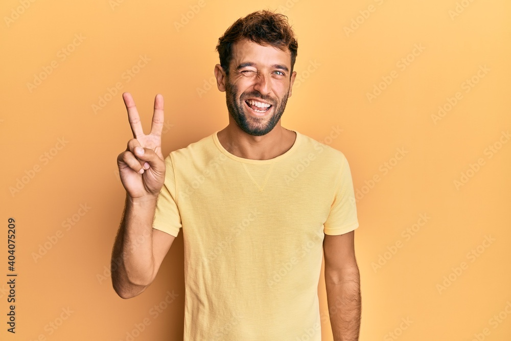 Handsome man with beard wearing casual yellow tshirt over yellow background smiling with happy face winking at the camera doing victory sign. number two. Handsome man with beard wearing casual yellow tshirt over yellow background smiling with happy face winking at the camera doing victory sign. number two.