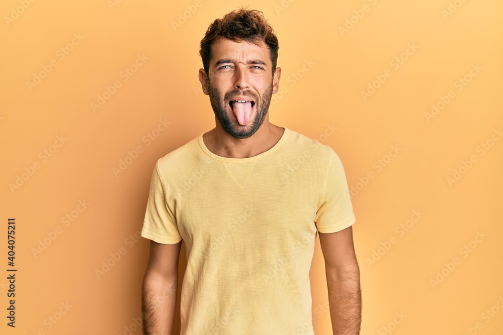 Handsome man with beard wearing casual yellow tshirt over yellow background sticking tongue out happy with funny expression. emotion concept. Handsome man with beard wearing casual yellow tshirt over yellow background sticking tongue out happy with funny expression. emotion concept.
