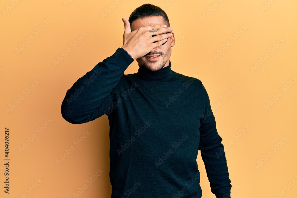 Handsome man with tattoos wearing turtleneck sweater and glasses smiling and laughing with hand on face covering eyes for surprise. blind concept. Handsome man with tattoos wearing turtleneck sweater and glasses smiling and laughing with hand on face covering eyes for surprise. blind concept.