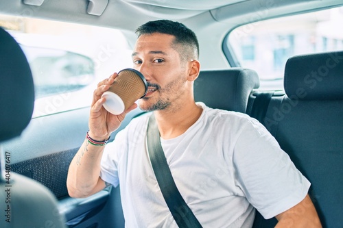 Young hispanic man smiling happy drinking take away coffee sitting on the car.