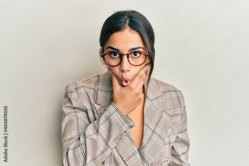 Young brunette woman wearing business jacket and glasses looking fascinated with disbelief, surprise and amazed expression with hands on chin