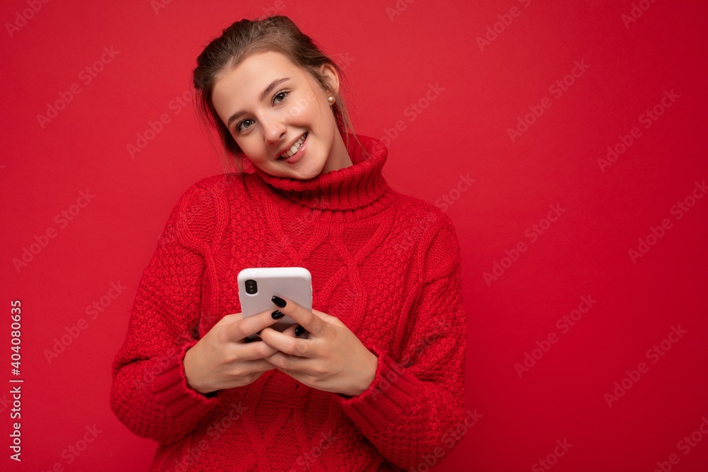 Attractive cute smiling young woman wearing warm red sweater isolated over red background wall holding smartphone and communicating online on the internet looking at camera Attractive cute smiling young woman wearing warm red sweater isolated over red background wall holding smartphone and communicating online on the internet looking at camera