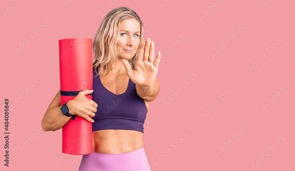 Middle age fit blonde woman holding yoga mat with open hand doing stop sign with serious and confident expression, defense gesture Middle age fit blonde woman holding yoga mat with open hand doing stop sign with serious and confident expression, defense gesture