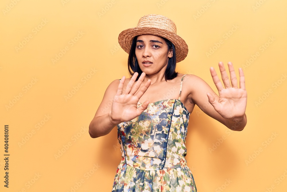 Young beautiful latin woman wearing summer hat afraid and terrified with fear expression stop gesture with hands, shouting in shock. panic concept. Young beautiful latin woman wearing summer hat afraid and terrified with fear expression stop gesture with hands, shouting in shock. panic concept.