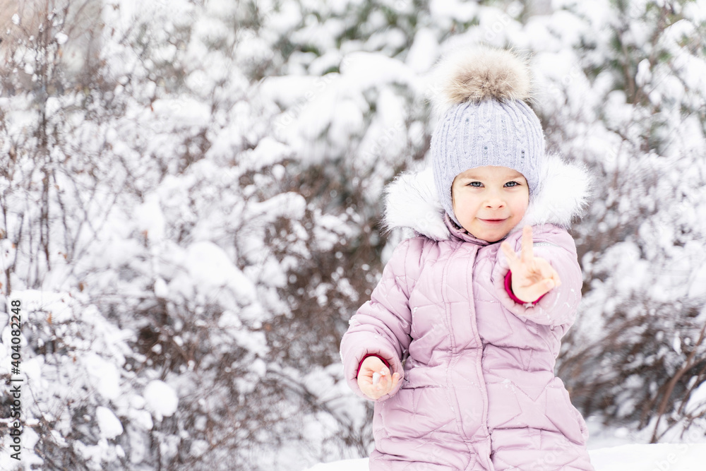 cute little girl in warm clothes and a hat in the winter forest. the child shows two fingers. copy space