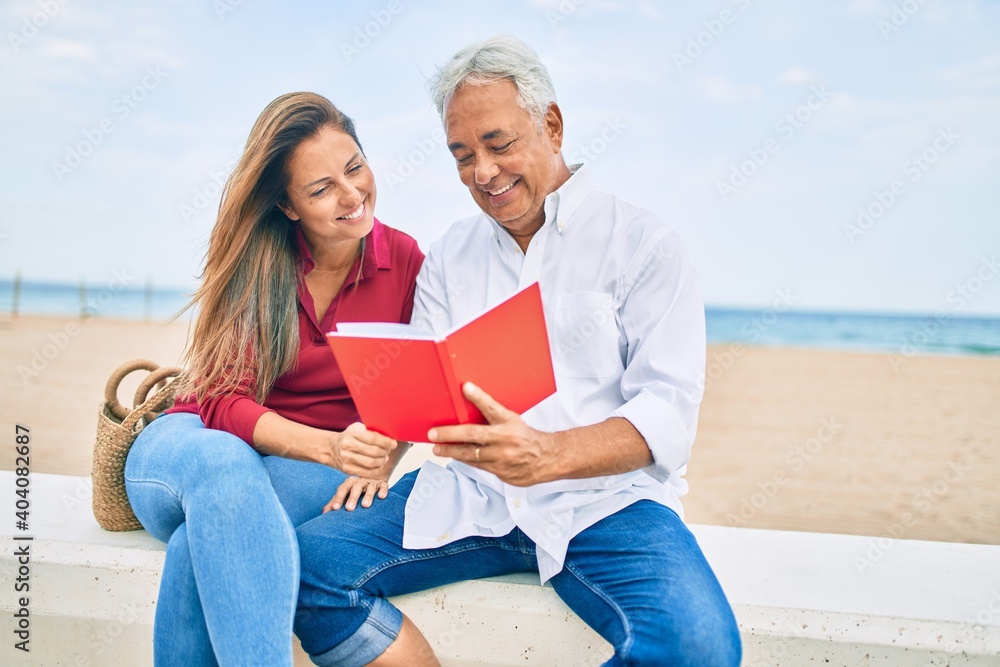 Middle age hispanic couple reading book sitting on the bench at the beach.