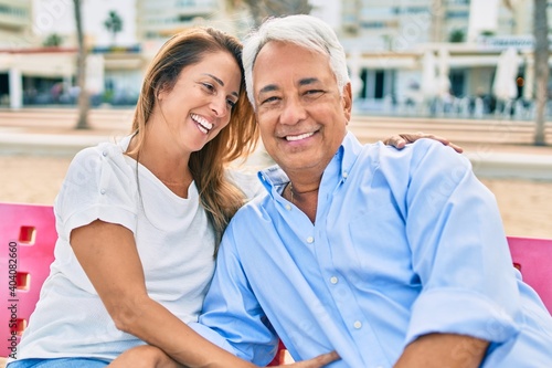 Middle age hispanic couple smiling happy and hugging sitting on bench at the beach.
