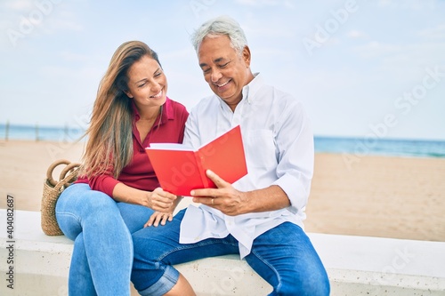 Middle age hispanic couple reading book sitting on the bench at the beach.