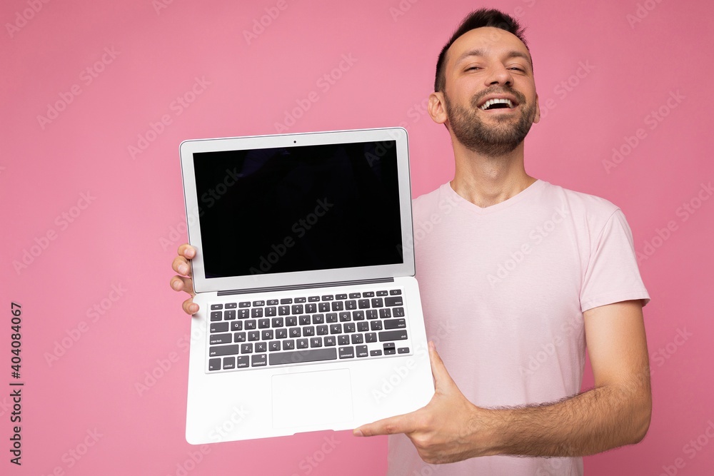 Handsome smiling brunet man holding laptop computer looking at camera in t-shirt on isolated pink background Handsome smiling brunet man holding laptop computer looking at camera in t-shirt on isolated pink background