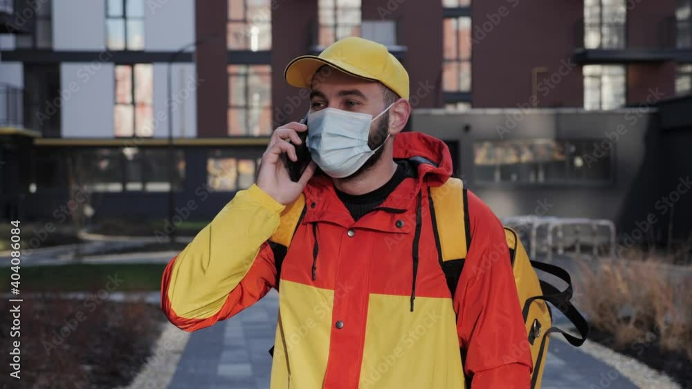 Attractive caucasian delivery man talks on the phone giving order in modern apartment complex with protective mask wears uniform and backpack with food. Sunny cold winter day isolation during epidemic Attractive caucasian delivery man talks on the phone giving order in modern apartment complex with protective mask wears uniform and backpack with food. Sunny cold winter day isolation during epidemic