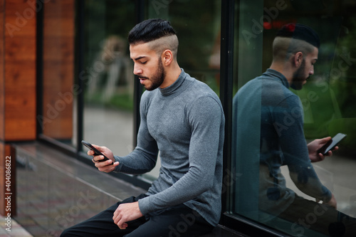 Portrait of stylish arab beard man wear grey turtleneck. Arabian model guy sit and look at smartphone.