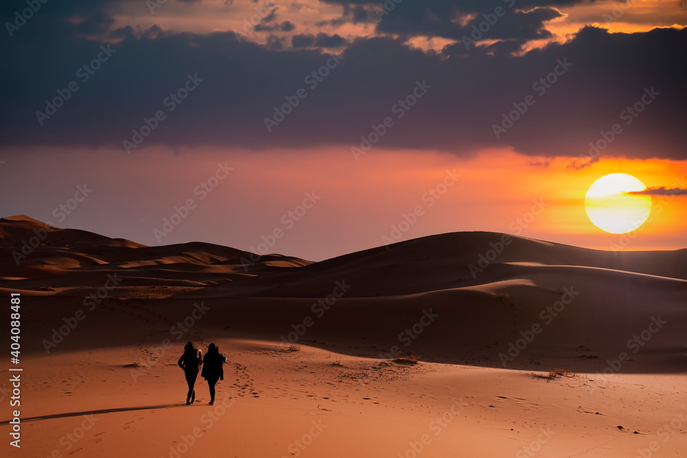 (Selective focus) Silhouette of two people walking on the sand dunes of the Merzouga desert during a stunning sunset. Merzouga, Morocco.