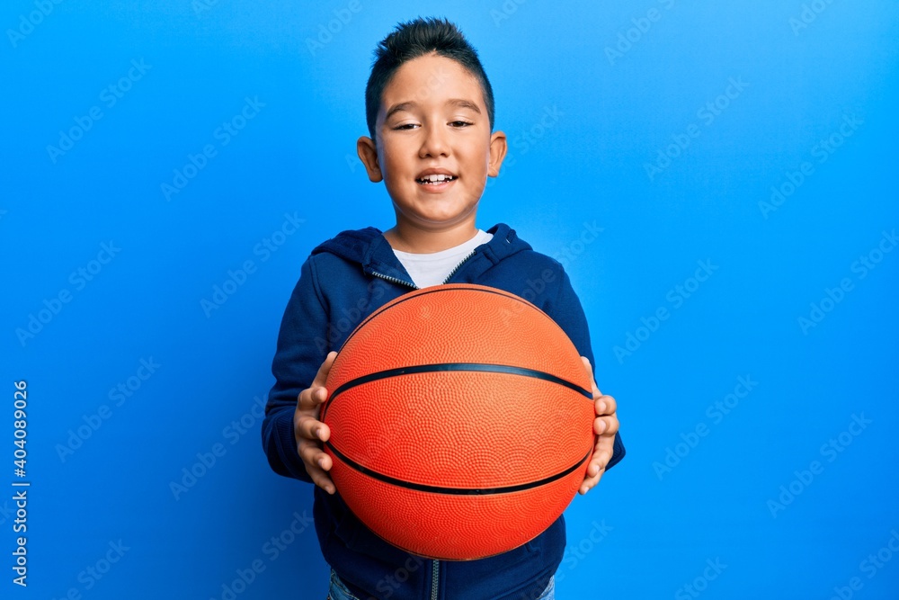 Little boy hispanic kid holding basketball ball winking looking at the camera with sexy expression, cheerful and happy face. Little boy hispanic kid holding basketball ball winking looking at the camera with sexy expression, cheerful and happy face.