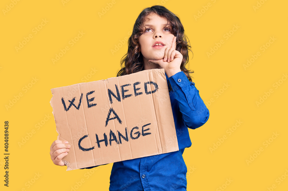 Cute hispanic child with long hair holding we need a change banner serious face thinking about question with hand on chin, thoughtful about confusing idea Cute hispanic child with long hair holding we need a change banner serious face thinking about question with hand on chin, thoughtful about confusing idea