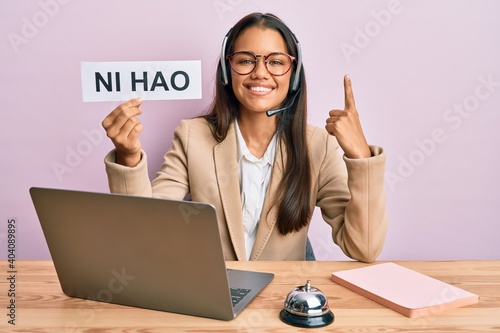 Beautiful hispanic woman wearing operator headset showing ni hao greeting smiling with an idea or question pointing finger with happy face, number one photo