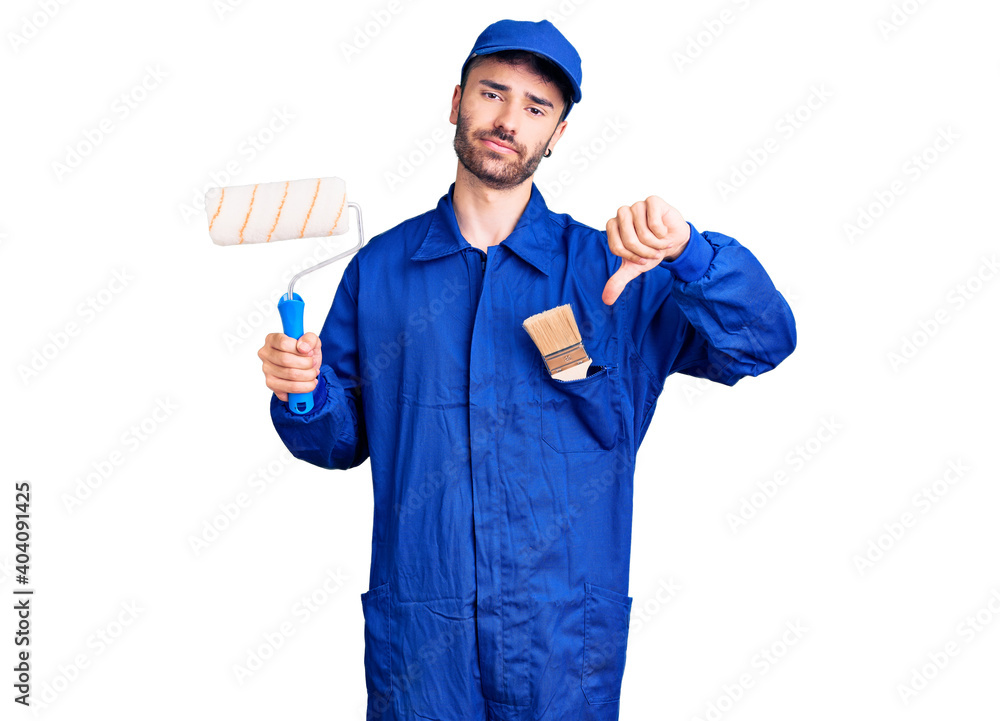 Young hispanic man wearing painter uniform holding roller with angry face, negative sign showing dislike with thumbs down, rejection concept