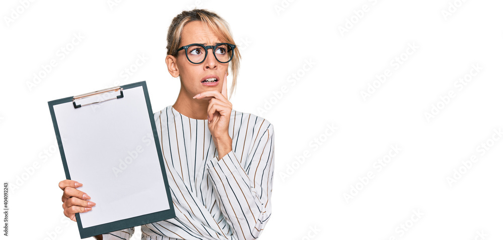 Beautiful blonde business woman holding clipboard with blank space serious face thinking about question with hand on chin, thoughtful about confusing idea Beautiful blonde business woman holding clipboard with blank space serious face thinking about question with hand on chin, thoughtful about confusing idea