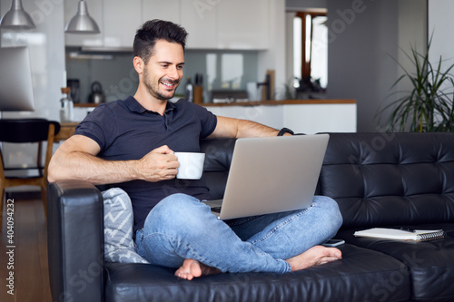 Relaxed man at home sitting with laptop on sofa drinking coffee