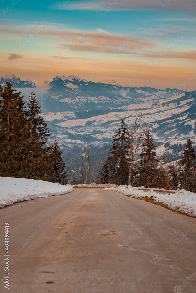Lonely asphalt road leading down towards the valley of Adelboden, looking towards Frutigen in winter time with some snow visible and early evening sunset time. Lonely asphalt road leading down towards the valley of Adelboden, looking towards Frutigen in winter time with some snow visible and early evening sunset time.