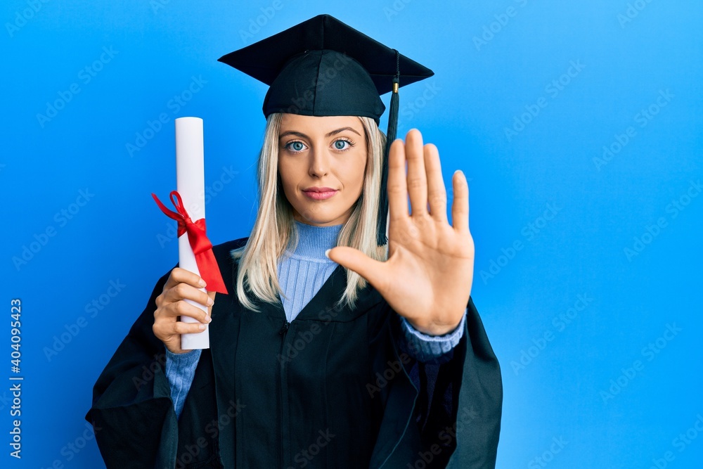 Beautiful blonde woman wearing graduation cap and ceremony robe holding degree with open hand doing stop sign with serious and confident expression, defense gesture Beautiful blonde woman wearing graduation cap and ceremony robe holding degree with open hand doing stop sign with serious and confident expression, defense gesture