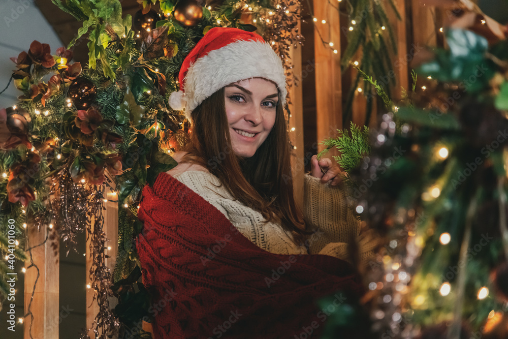 Portrait of beautiful caucasian woman in Santa hat sitting on wooden staircase decorated for Christmas and New Year. Christmas home celebration theme. Portrait of beautiful caucasian woman in Santa hat sitting on wooden staircase decorated for Christmas and New Year. Christmas home celebration theme.