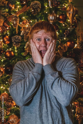 Portrait of shocked caucasian man at Christmas. Man with open mouth holds his face like in the movie Home Alone. Blurred Christmas tree in the background. Holiday emotions theme. photo