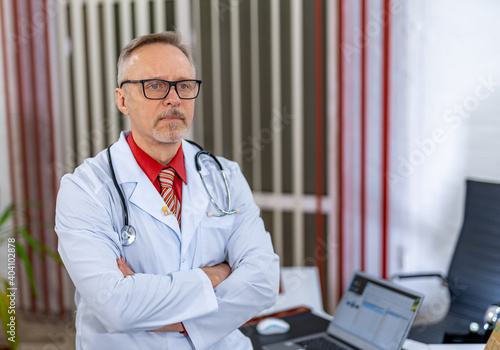 Doctor stands crosshands in medical office. Modern clinic background. Doctor in glasses in scrubs and stethoscope on neck. photo