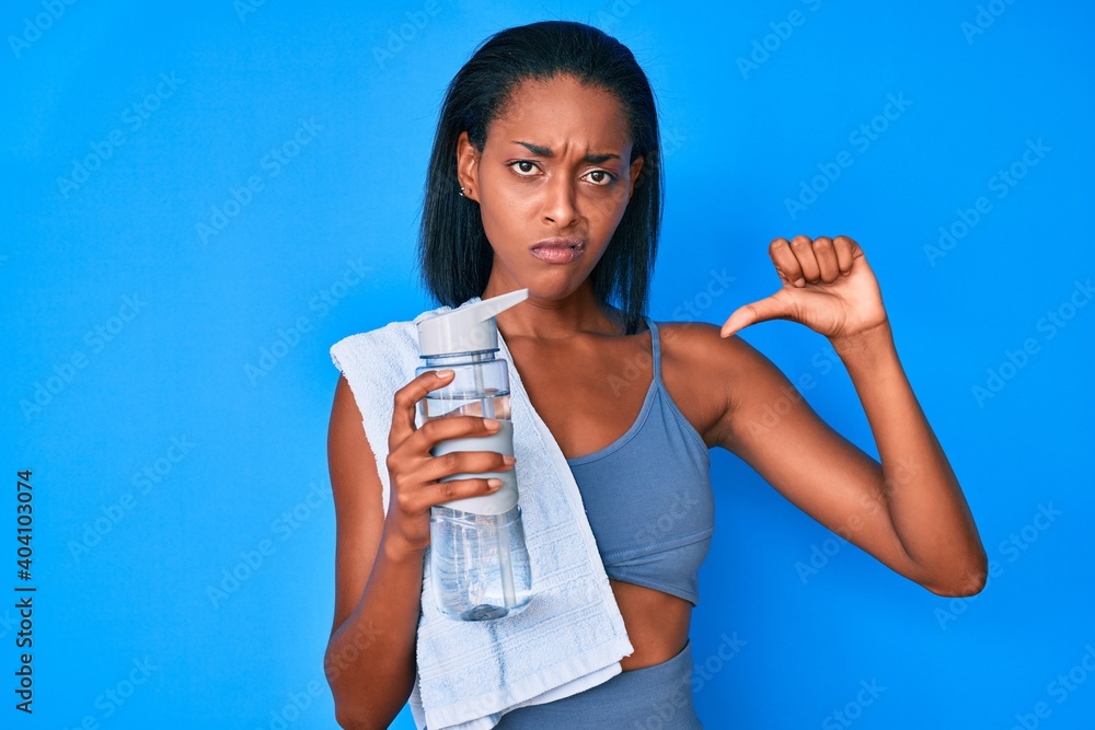 Young african american woman wearing sportswear drinking bottle of water with angry face, negative sign showing dislike with thumbs down, rejection concept Young african american woman wearing sportswear drinking bottle of water with angry face, negative sign showing dislike with thumbs down, rejection concept