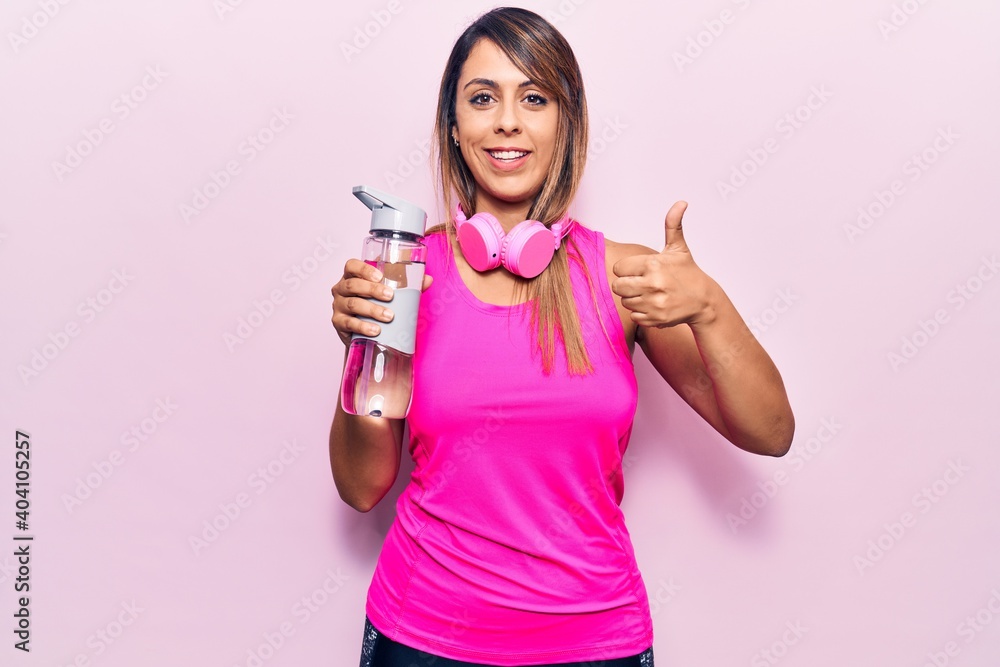 Young beautiful woman wearing sportswear drinking bottle of water using headphones smiling happy and positive, thumb up doing excellent and approval sign Young beautiful woman wearing sportswear drinking bottle of water using headphones smiling happy and positive, thumb up doing excellent and approval sign