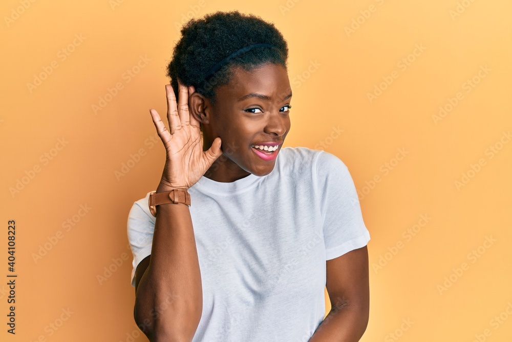 Young african american girl wearing casual white t shirt smiling with hand over ear listening and hearing to rumor or gossip. deafness concept. Young african american girl wearing casual white t shirt smiling with hand over ear listening and hearing to rumor or gossip. deafness concept.