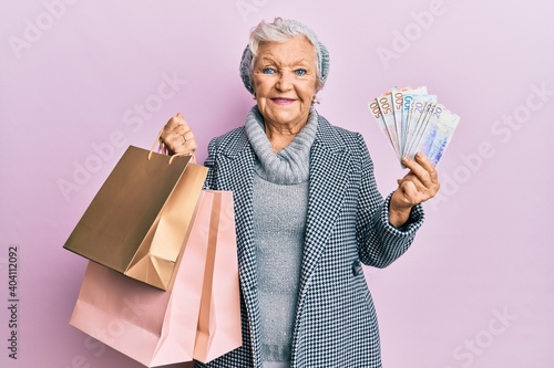 Senior grey-haired woman holding shopping bags and swedish krona banknotes smiling with a happy and cool smile on face. showing teeth.