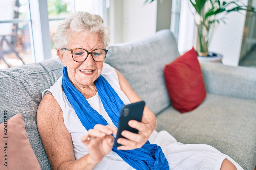Elder senior woman with grey hair smiling happy sitting on the sofa at home using smartphone Elder senior woman with grey hair smiling happy sitting on the sofa at home using smartphone