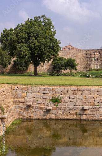 Hampi, Karnataka, India - November 5, 2013: Zanana Enclosure. Corner of borwn stone Queens pool or tank with green foliage and protection wall in back under light blue cloudscape. Scaffold in back. photo