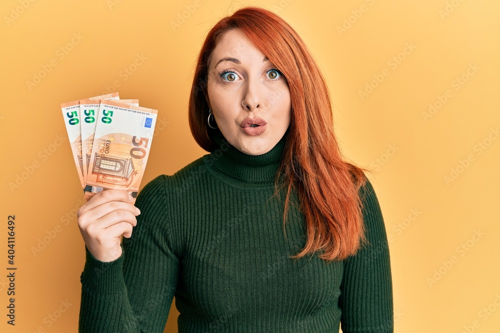 Beautiful redhead woman holding bunch of 50 euro banknotes scared and amazed with open mouth for surprise, disbelief face Beautiful redhead woman holding bunch of 50 euro banknotes scared and amazed with open mouth for surprise, disbelief face
