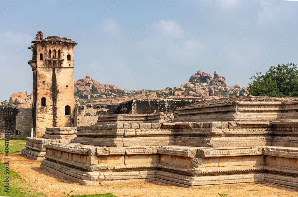 Hampi, Karnataka, India - November 5, 2013: Zanana Enclosure. Remains of Queens Palace is base platform. Brown stone watchtower in back under blue sky. Mountains and green foliage as backdrop. Hampi, Karnataka, India - November 5, 2013: Zanana Enclosure. Remains of Queens Palace is base platform. Brown stone watchtower in back under blue sky. Mountains and green foliage as backdrop.
