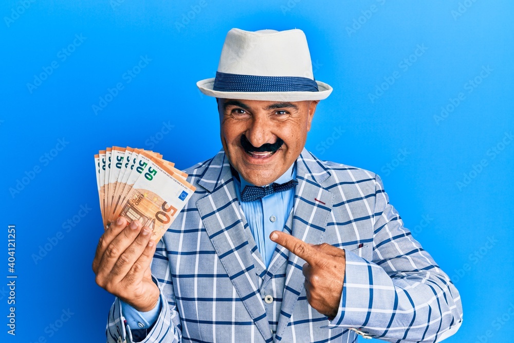 Mature middle east man with mustache wearing elegant vintage style holding euros banknotes smiling happy pointing with hand and finger Mature middle east man with mustache wearing elegant vintage style holding euros banknotes smiling happy pointing with hand and finger