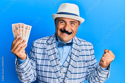 Mature middle east man with mustache wearing elegant vintage style holding pounds banknotes screaming proud, celebrating victory and success very excited with raised arm photo