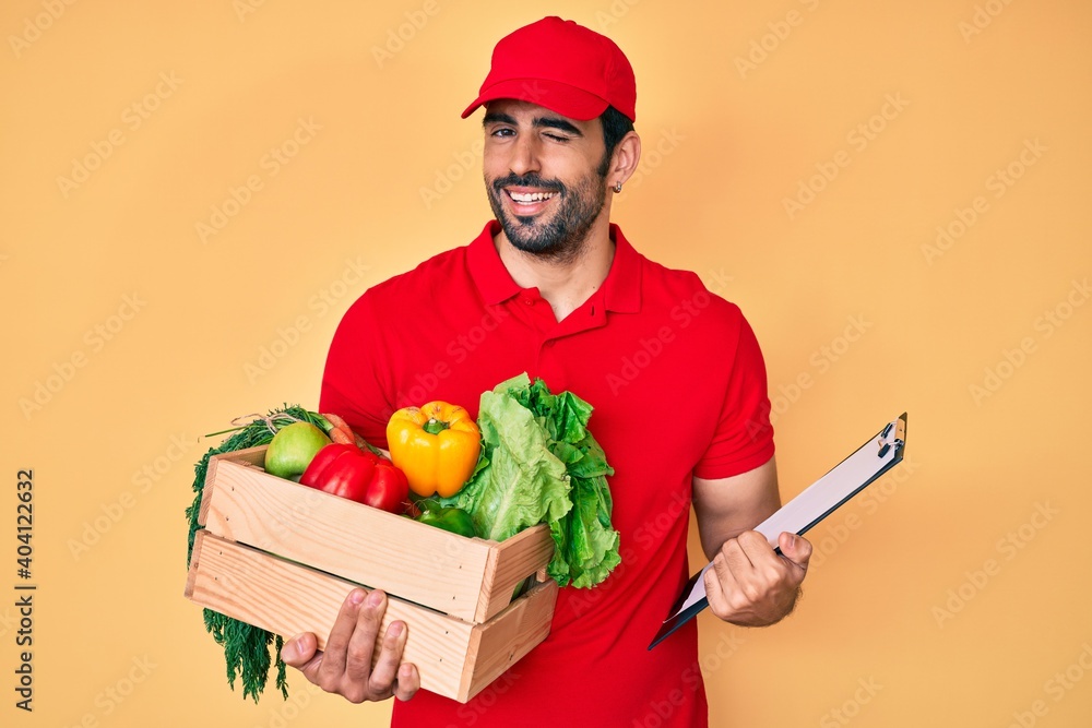Handsome hispanic man with beard wearing uniform holding delivery food winking looking at the camera with sexy expression, cheerful and happy face. Handsome hispanic man with beard wearing uniform holding delivery food winking looking at the camera with sexy expression, cheerful and happy face.