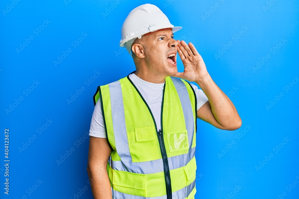 Middle age bald man wearing architect hardhat shouting and screaming loud to side with hand on mouth. communication concept.