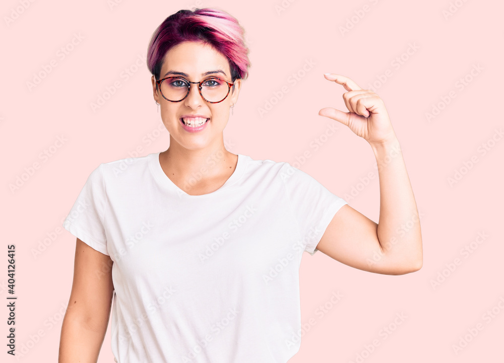 Young beautiful woman with pink hair wearing casual clothes and glasses smiling and confident gesturing with hand doing small size sign with fingers looking and the camera. measure concept.