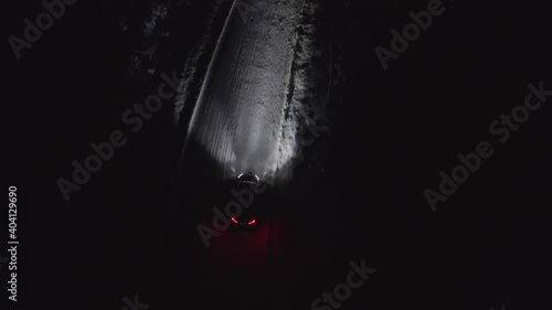 Car or vehicle driving on a snowy road in the winter at night. Headlights lighting up the icy road in the dark. Heading away to distance. Following or tracking aerial drone shot.