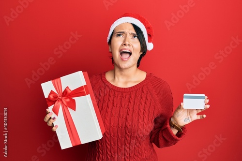 Beautiful young woman with short hair wearing chrismast hat holding gift and credit card angry and mad screaming frustrated and furious, shouting with anger looking up. photo