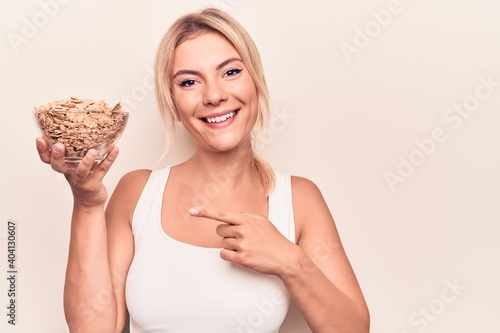 Young beautiful blonde woman holding bowl with conrflakes cerals over white background smiling happy pointing with hand and finger