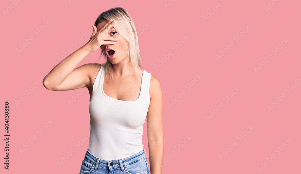 Young beautiful blonde woman wearing casual sleeveless t-shirt peeking in shock covering face and eyes with hand, looking through fingers with embarrassed expression. Young beautiful blonde woman wearing casual sleeveless t-shirt peeking in shock covering face and eyes with hand, looking through fingers with embarrassed expression.