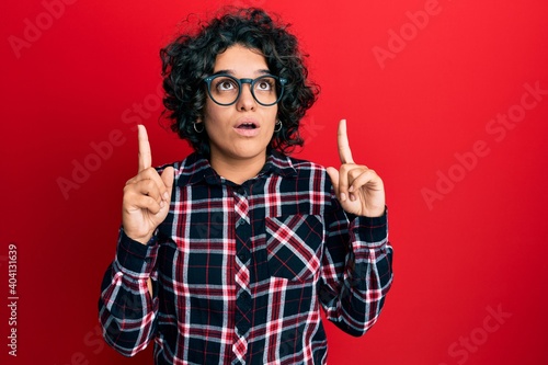 Young hispanic woman with curly hair wearing casual clothes and glasses amazed and surprised looking up and pointing with fingers and raised arms.