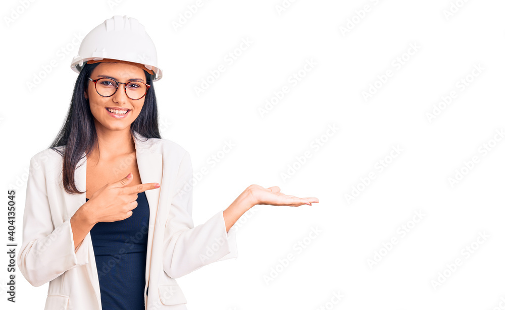 Young beautiful latin girl wearing architect hardhat and glasses amazed and smiling to the camera while presenting with hand and pointing with finger. Young beautiful latin girl wearing architect hardhat and glasses amazed and smiling to the camera while presenting with hand and pointing with finger.