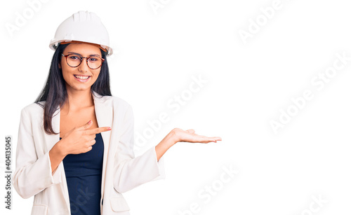 Young beautiful latin girl wearing architect hardhat and glasses amazed and smiling to the camera while presenting with hand and pointing with finger.