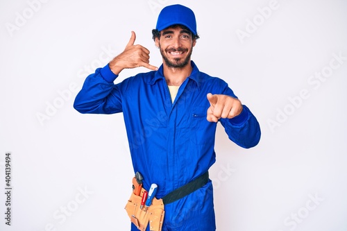 Handsome young man with curly hair and bear weaing handyman uniform smiling doing talking on the telephone gesture and pointing to you. call me.