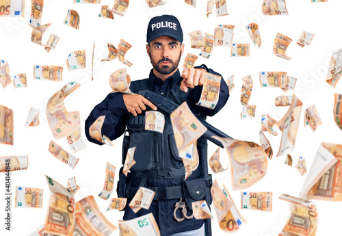 Young hispanic man wearing police uniform holding shotgun pointing with finger to the camera and to you, confident gesture looking serious