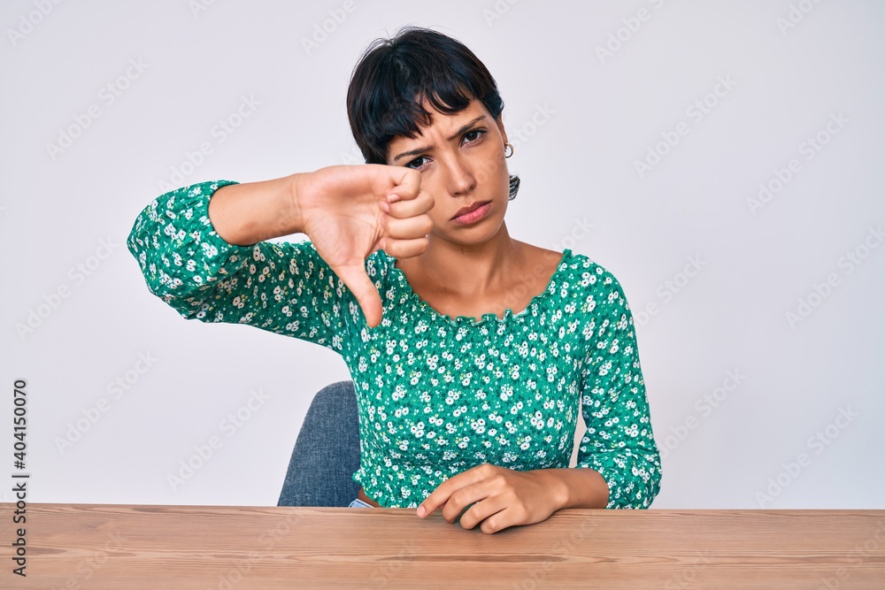 Beautiful brunettte woman wearing casual clothes sitting on the table looking unhappy and angry showing rejection and negative with thumbs down gesture. bad expression. Beautiful brunettte woman wearing casual clothes sitting on the table looking unhappy and angry showing rejection and negative with thumbs down gesture. bad expression.